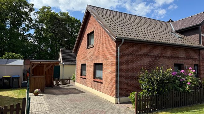 Außenansicht eines roten Backsteinhauses mit Satteldach, Einfahrt, Gartenzaun und blühenden Sträuchern.