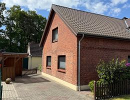Außenansicht eines roten Backsteinhauses mit Satteldach, Einfahrt, Gartenzaun und blühenden Sträuchern.